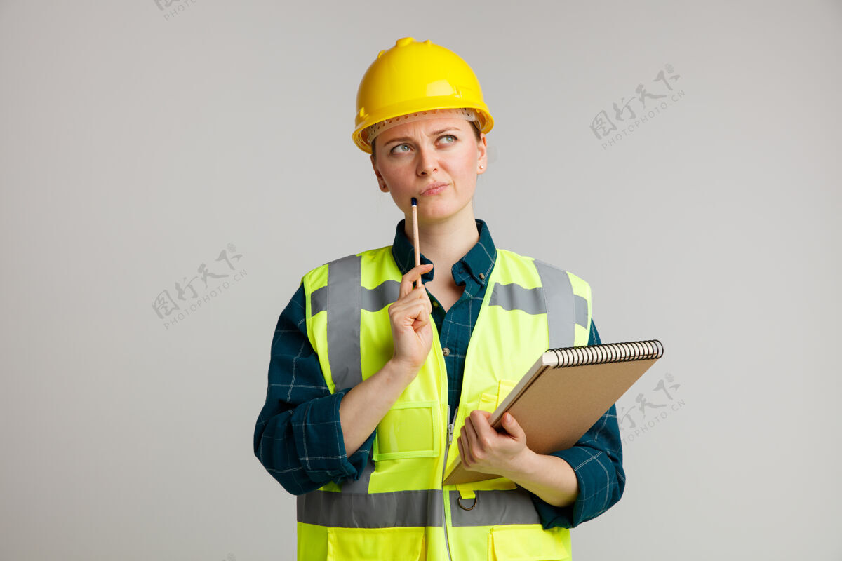 怀疑可疑的年轻女建筑工人戴着安全帽和安全背心拿着记事本用铅笔摸下巴抬头看年轻人头盔背心
