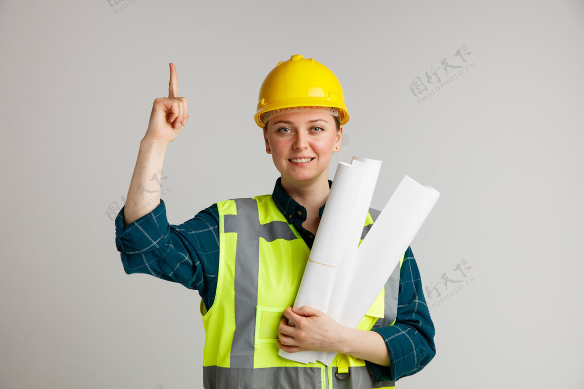 头盔面带微笑的年轻女建筑工人戴着安全帽和安全背心 拿着纸指着上白色年轻戴着