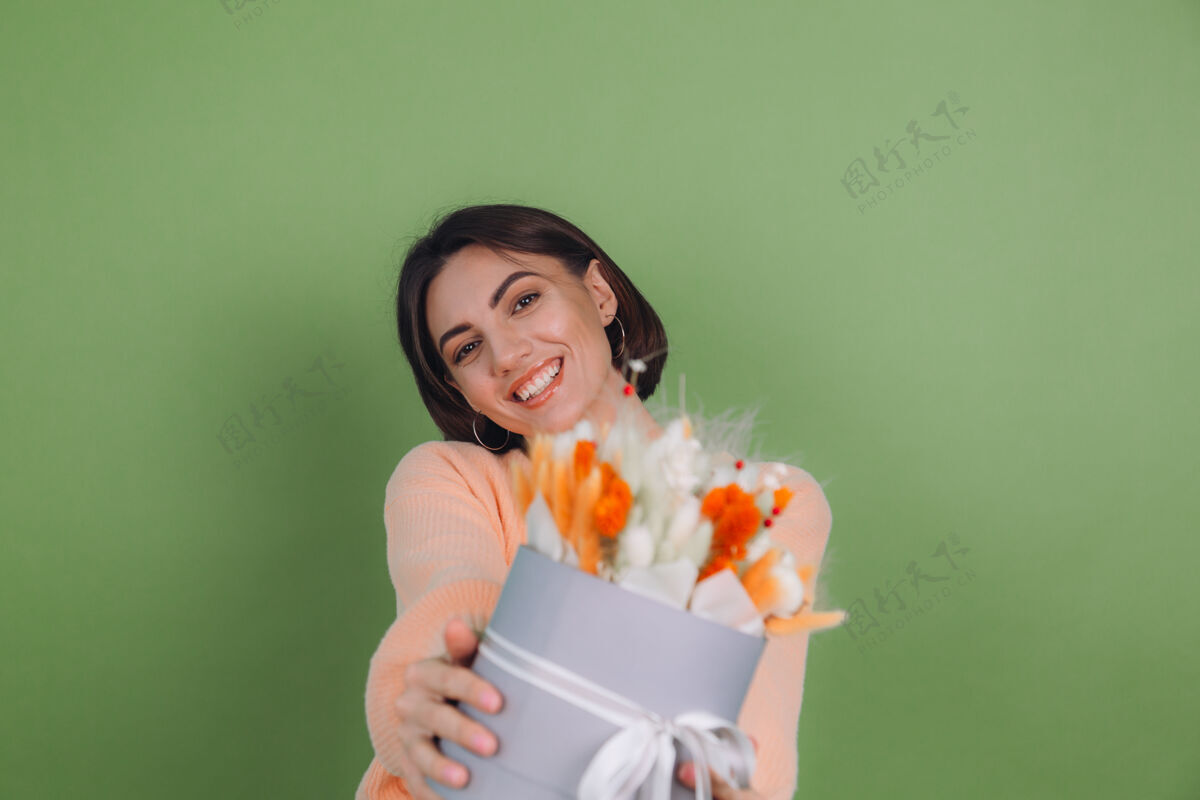 棉花花年轻女子穿着休闲桃色毛衣隔离在绿橄榄墙上手持橙白色花盒组成的棉花 吉普赛拉小麦和拉古鲁斯作为礼物开心惊喜年轻积极蝴蝶结