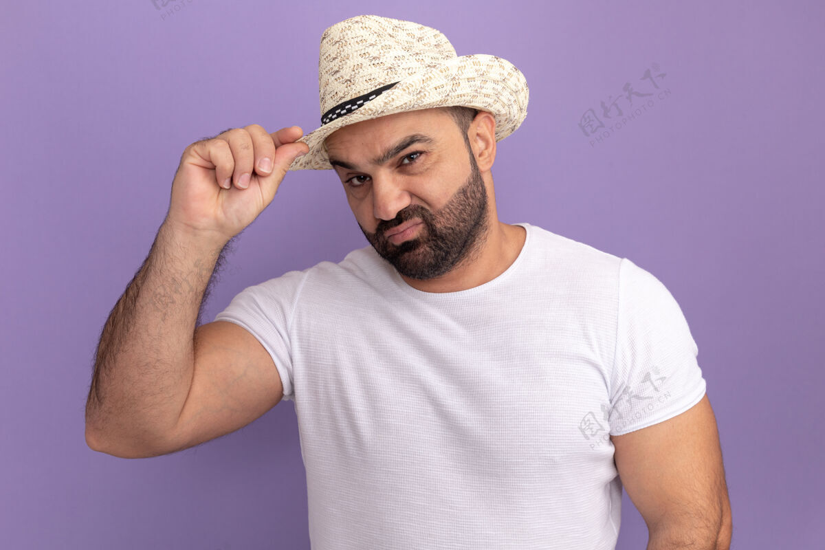 男人一个留着胡子的男人 穿着白色t恤 戴着夏令帽 脸上带着怀疑的表情 站在紫色的墙上摸着他的帽子立场胡须帽子