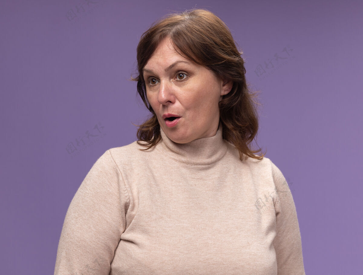 高领毛衣穿着米色套头衫的中年妇女站在紫色的墙上 困惑而惊讶地向一旁望去站中年年龄
