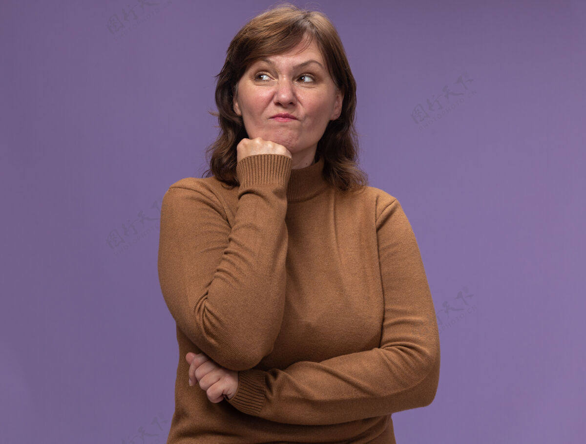 旁边不满意的中年妇女穿着棕色高领毛衣 手放在下巴上 一边看着一边 一边想着站在紫色的墙上女人思考中间
