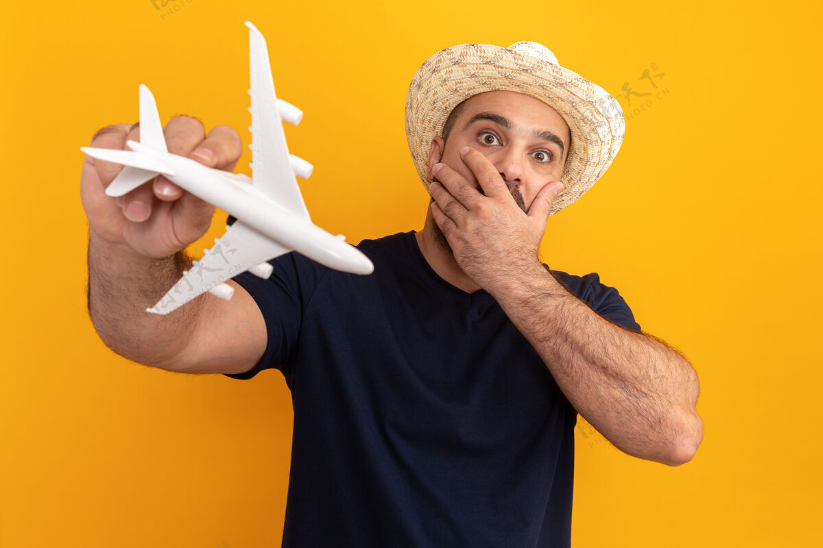 飞机留着胡子的男人穿着黑色t恤 戴着夏天的帽子 手里拿着玩具飞机 惊讶又担心地捂着嘴 手站在橙色的墙上胡须手势忧虑