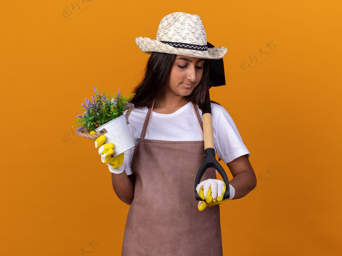 花园年轻的园丁女孩拿着盆栽和铲子站在橙色的墙上 看起来很自信锅年轻铲