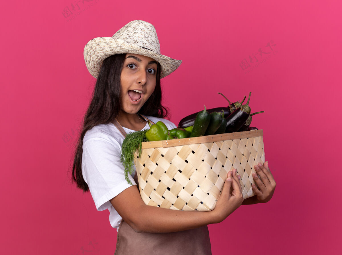 欢呼穿着围裙 戴着夏帽 手里拿着装满蔬菜的箱子的年轻园丁女孩站在粉红色的墙上 高兴而兴奋地微笑着兴奋板条箱围裙