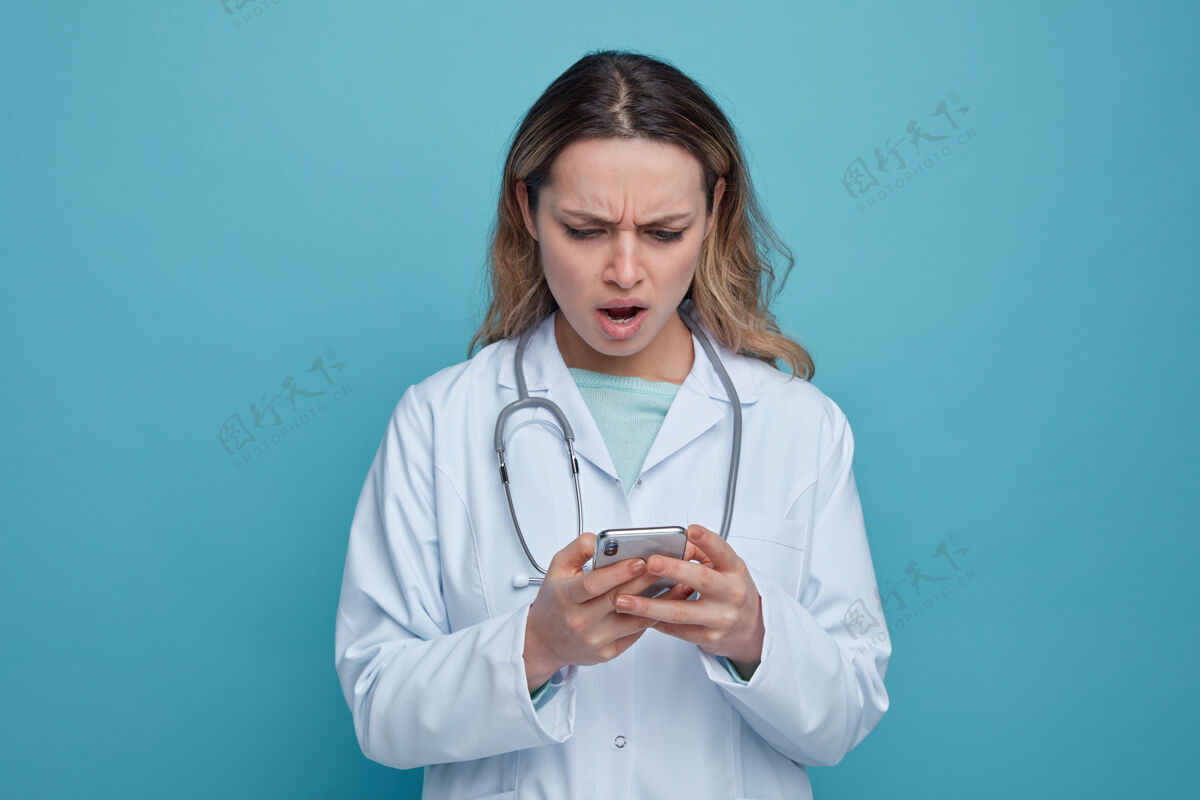 长袍恼火的年轻女医生穿着医用长袍 脖子上戴着听诊器用手机使用年轻医疗