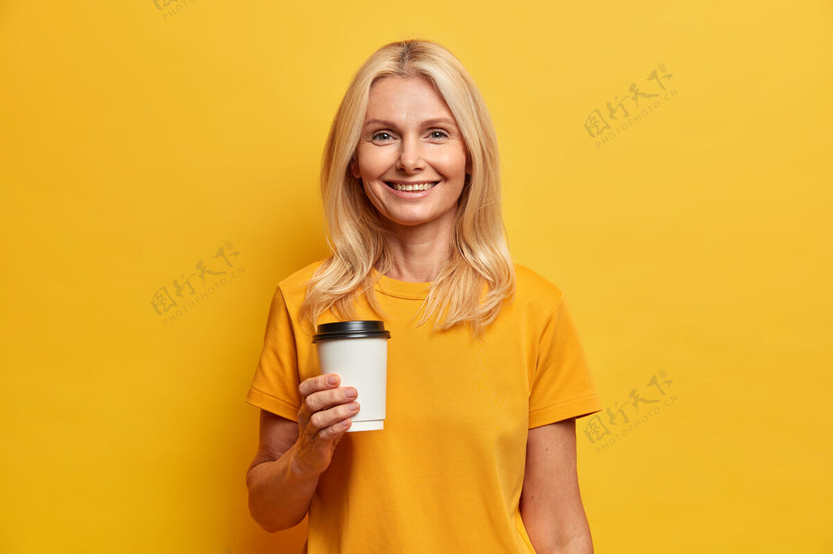 女性欧洲金发女人的水平镜头 带着愉快的微笑 化着最少的妆 手里拿着一杯咖啡 穿着休闲t恤请外卖积极