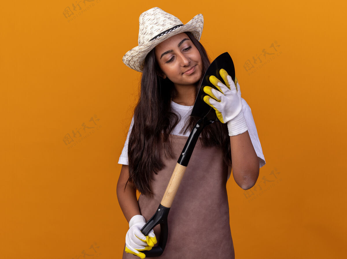 帽子年轻的园丁女孩围着围裙 戴着夏帽 拿着铲子 站在橙色的墙上 面带微笑地看着它站着花园围裙