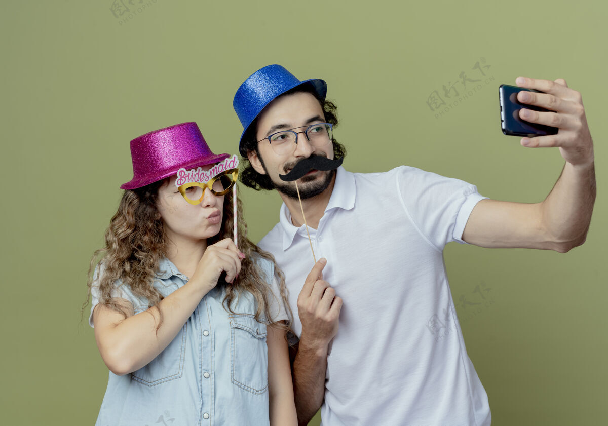 假一对戴着粉蓝相间帽子的年轻夫妇自拍了一张照片 照片上的女孩戴着化装眼罩 男孩戴着假胡子粉色抱着棍子