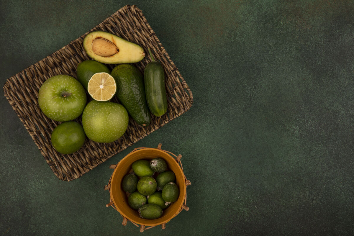 营养俯视图：绿色表面上的柳条托盘上的绿色苹果 鳄梨 黄瓜桶上的feijoas饮食健康水桶