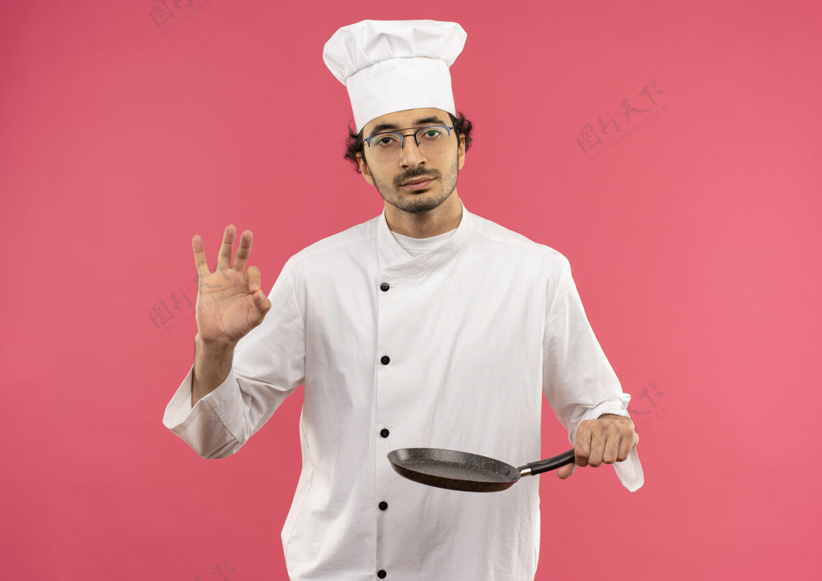 年轻自信的年轻男厨师穿着厨师制服 戴着眼镜 手里拿着煎锅 摆出一副很好的姿势粉红男拿着