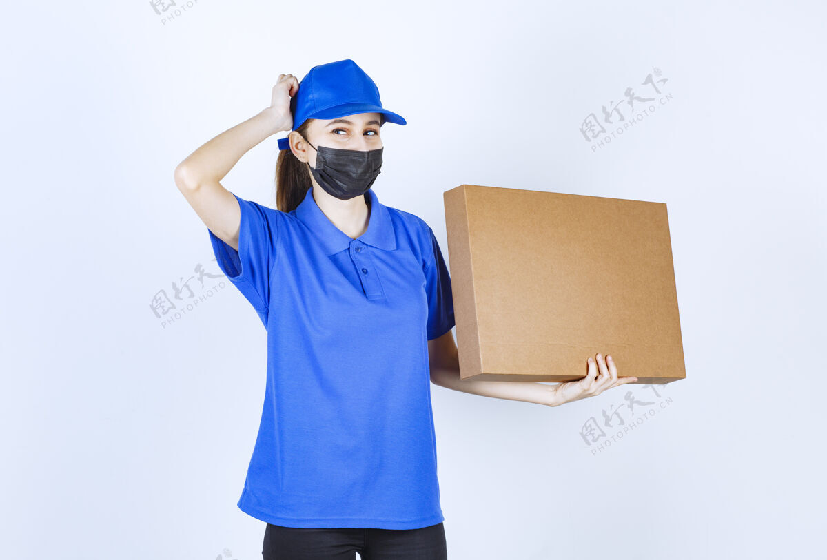思想戴着面具 穿着蓝色制服的女快递员手里拿着一个大的纸板包裹 看上去困惑而犹豫不确定外卖服装