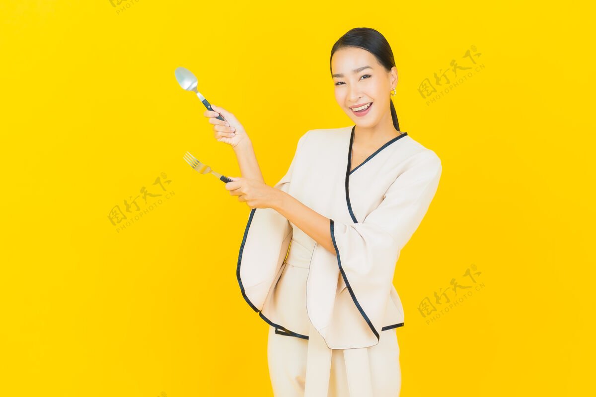 手在黄色的墙上 一位年轻的亚洲商界美女拿着勺子和叉子准备吃东西准备叉子盘子