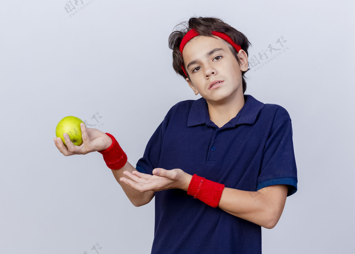 运动笨手笨脚的年轻帅气的运动男孩戴着头带和带牙套的腕带手拿着并指着隔离在白墙上的苹果穿牙科头带