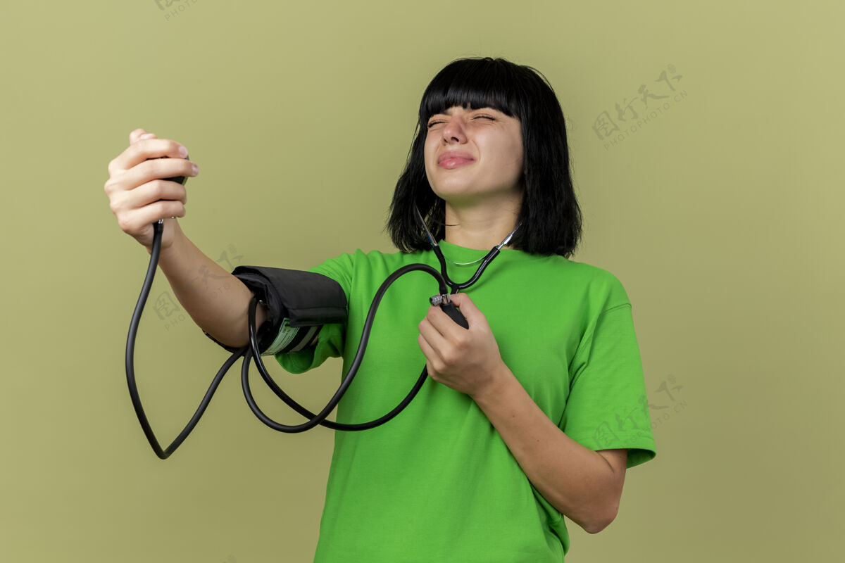 佩戴焦虑的年轻白种人女孩戴着听诊器 闭着眼睛用血压计测量血压 隔离在橄榄绿的墙上 留有复印空间压力封闭年轻
