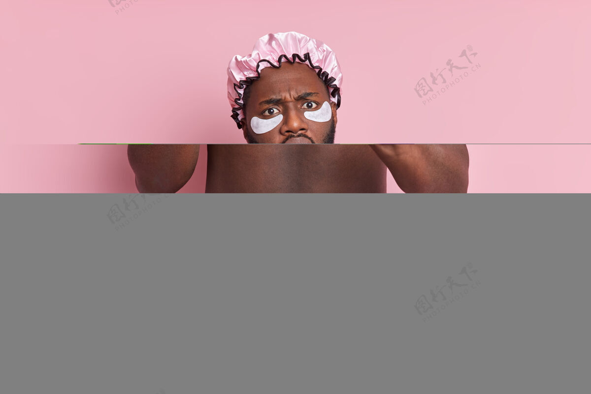 洗澡迷惑不解的男人保持手掌向前 淋浴时拿着海绵 戴着防水帽 眼睛下面贴着美容贴片 对着粉色的墙壁摆姿势男人反应手掌