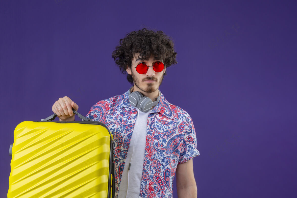 男人年轻帅气的卷发旅行者戴着太阳镜和耳机 脖子上拿着手提箱 在隔离的紫色墙壁上看着右边有复印空间耳机帅气侧身