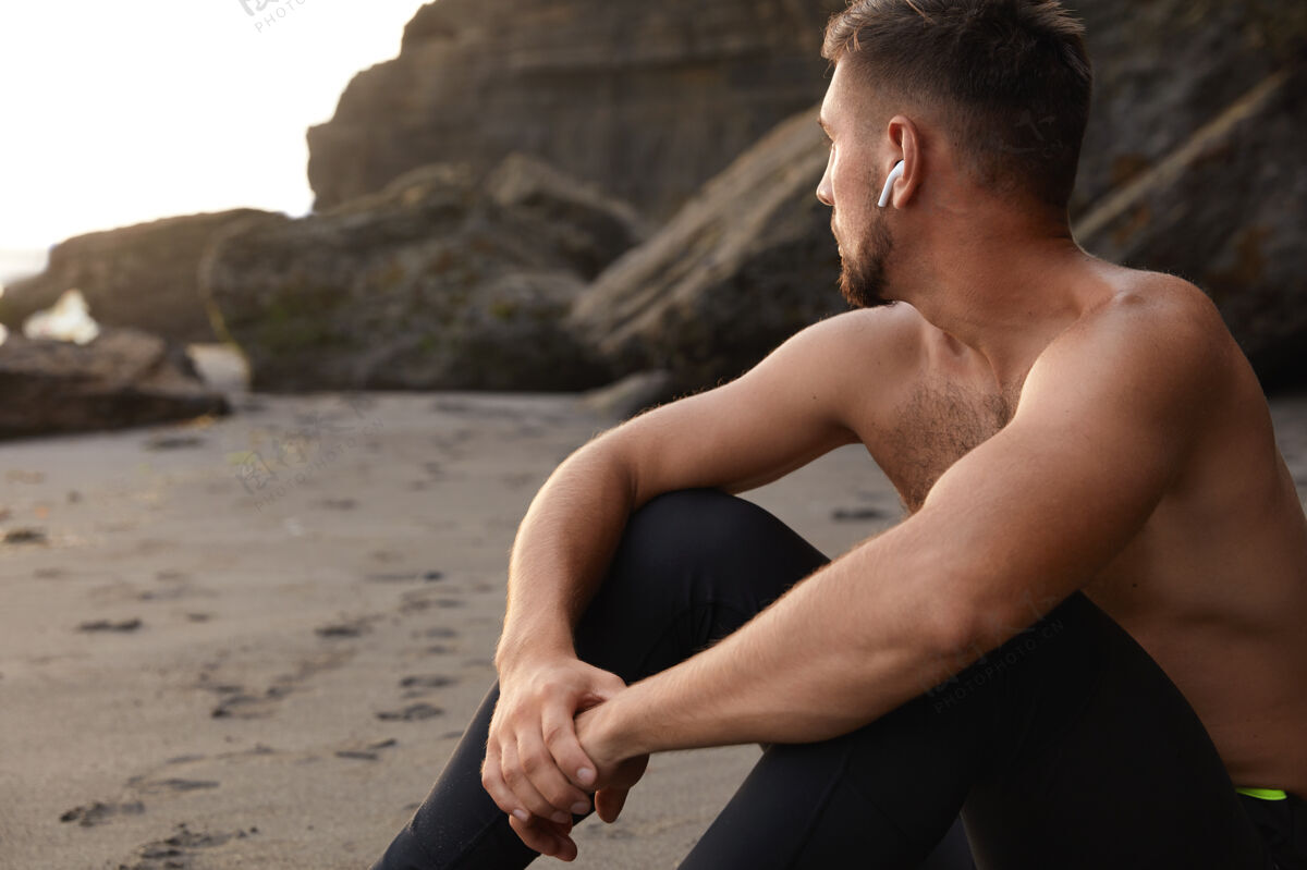 运动服沉思的运动员坐在沙滩上的水平视图 聚焦在远处跑步音频专注