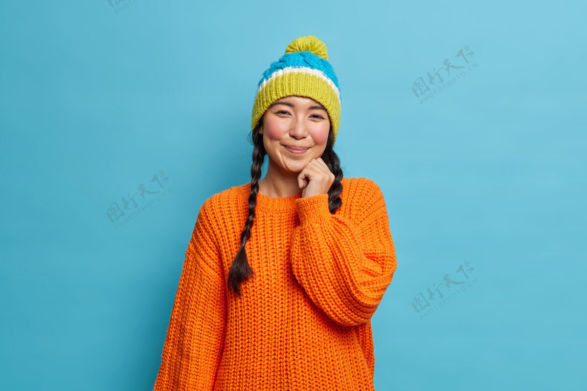 欢呼一个漂亮害羞的深色头发的亚洲女人的肖像 留着两条辫子 穿着针织毛衣 戴着帽子 有着满意的面部表情 在工作室里对着蓝色的墙壁在冬天散步毛衣节日美女