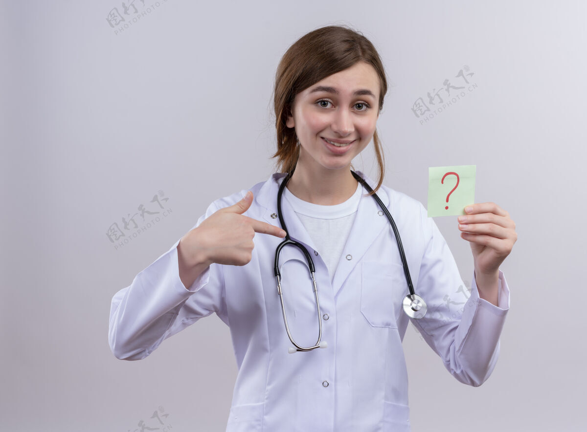 年轻人面带微笑的年轻女医生 穿着医用长袍 戴着听诊器 对着写在纸上的问号 贴在隔离的白色墙上听诊器问题医生