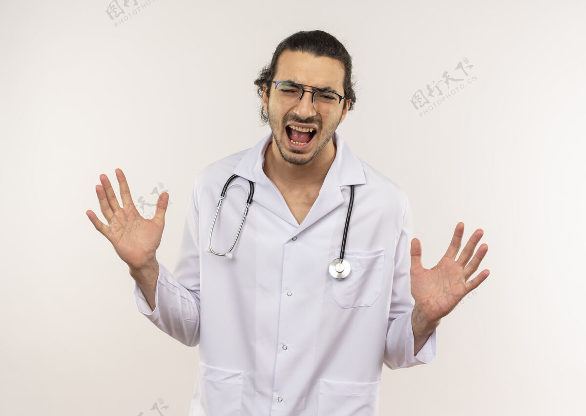 传播愤怒的年轻男医生 戴着眼镜 穿着白色长袍 戴着听诊器 闭着眼睛 把手放在隔离的白色墙壁上 留着复印空间穿封闭长袍