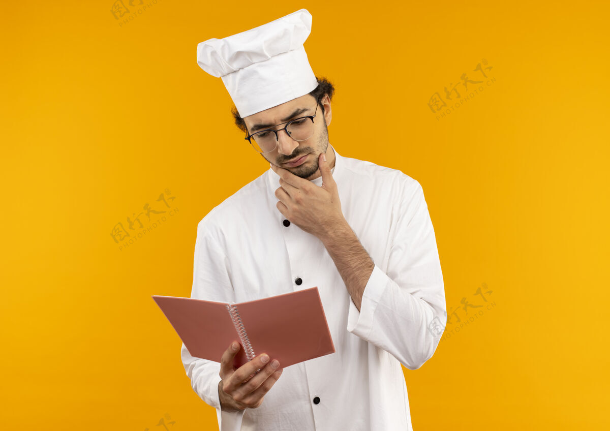 手想着年轻的男厨师穿着厨师制服 戴着眼镜 拿着笔记本看着 把手放在隔离在黄色墙上的下巴上烹饪持有男性