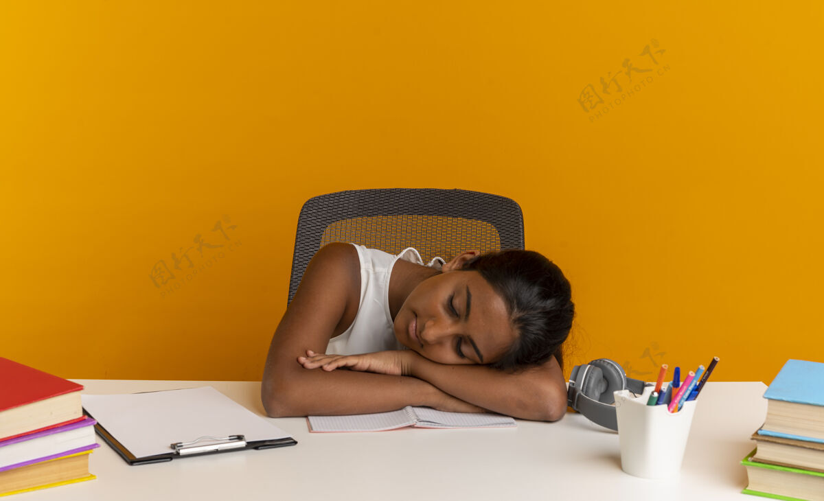 关门年轻的女学生闭着眼睛坐在课桌旁 手里拿着学习用具 把头靠在桌子上 被隔离在橘色的墙上头年轻女学生