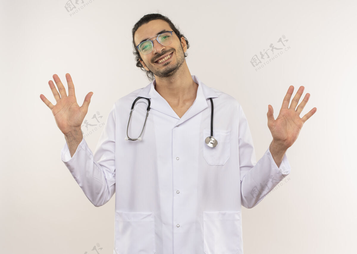 男性微笑着的年轻男医生戴着眼镜 穿着白色长袍 带着听诊器 在隔离的白色墙壁上摊开双手 留着复印空间年轻光学传播