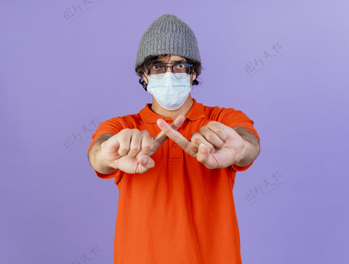 穿着年轻的白种人 戴着眼镜 戴着口罩 戴着冬天的帽子 不做任何手势 孤零零地站在紫色的墙上 留着复制的空间姿势年轻人疾病