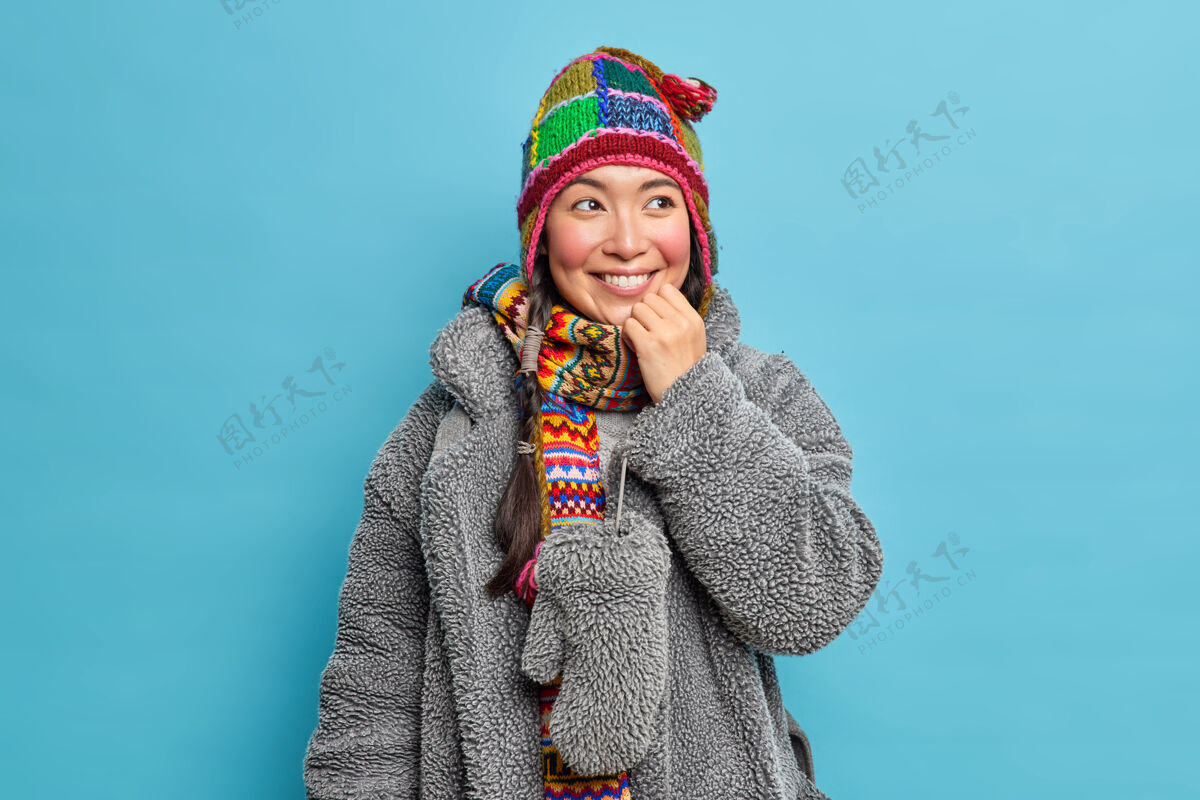 外套高兴的亚洲女人戴着针织帽子围巾和灰色外套从天然皮毛微笑积极有好心情隔着蓝色的墙壁衣服女性围巾