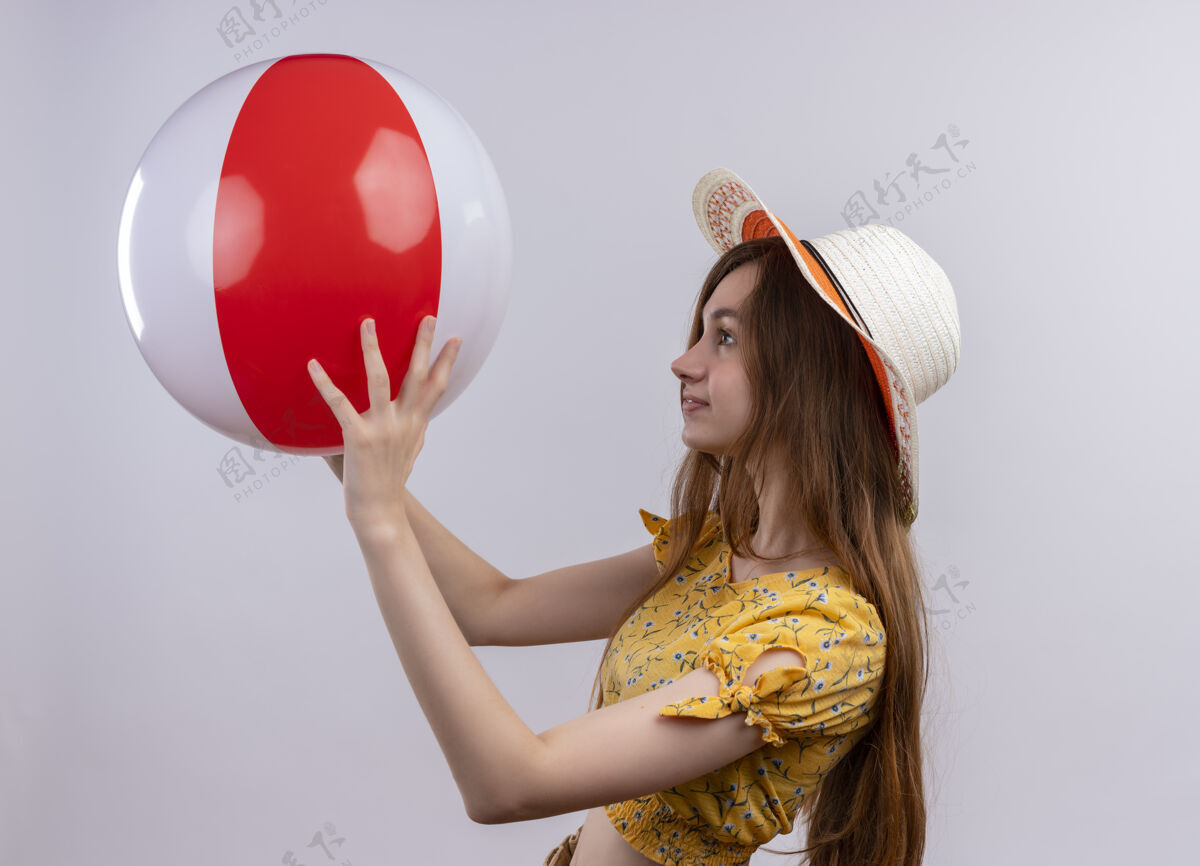 女人戴着帽子的年轻女孩举起沙滩球 站在隔离的白色墙壁上 看着它的侧视图年轻人提高看