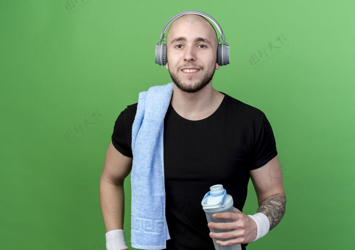 腕带微笑的年轻人戴着护腕和耳机 肩上扛着毛巾的水瓶 隔离在绿色的墙上毛巾男人瓶子