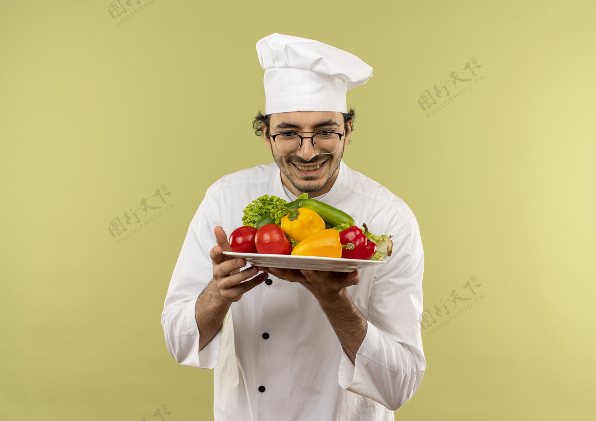 男性面带微笑的年轻男厨师穿着厨师制服 戴着眼镜 拿着蔬菜看着隔离在绿墙上的盘子微笑蔬菜盘子