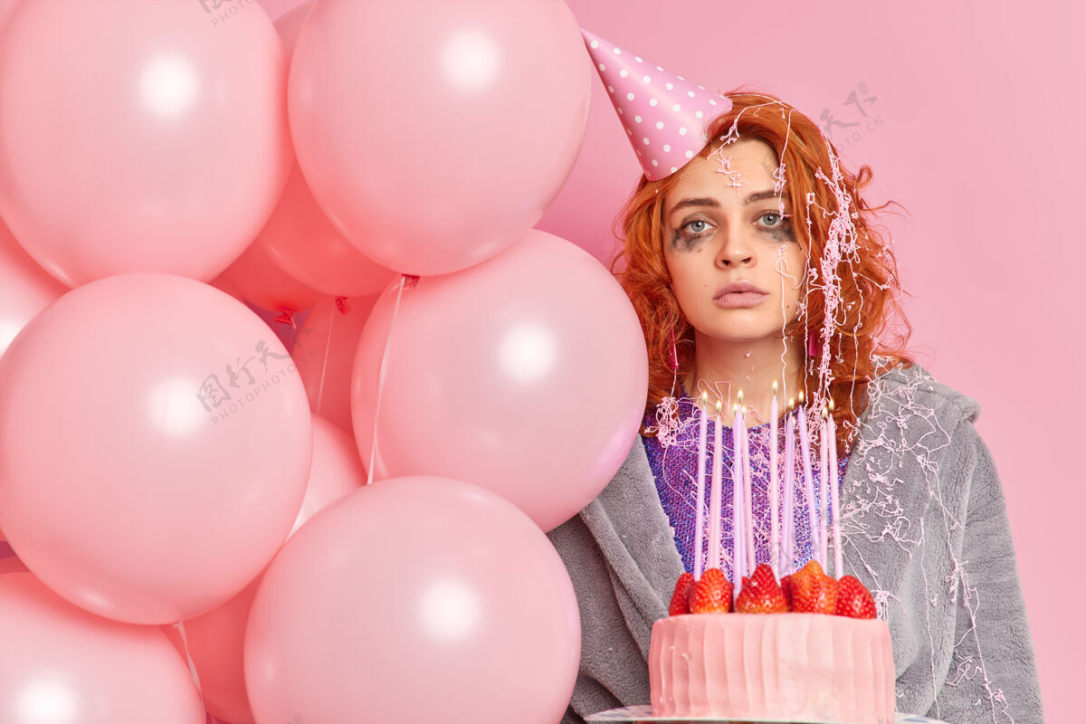 氦严肃的红发女人 漏了妆 直视前方 戴着派对锥形帽 睡衣 拿着美味的草莓蛋糕 充气气球 对着粉色的墙壁摆姿势聚会姜脸