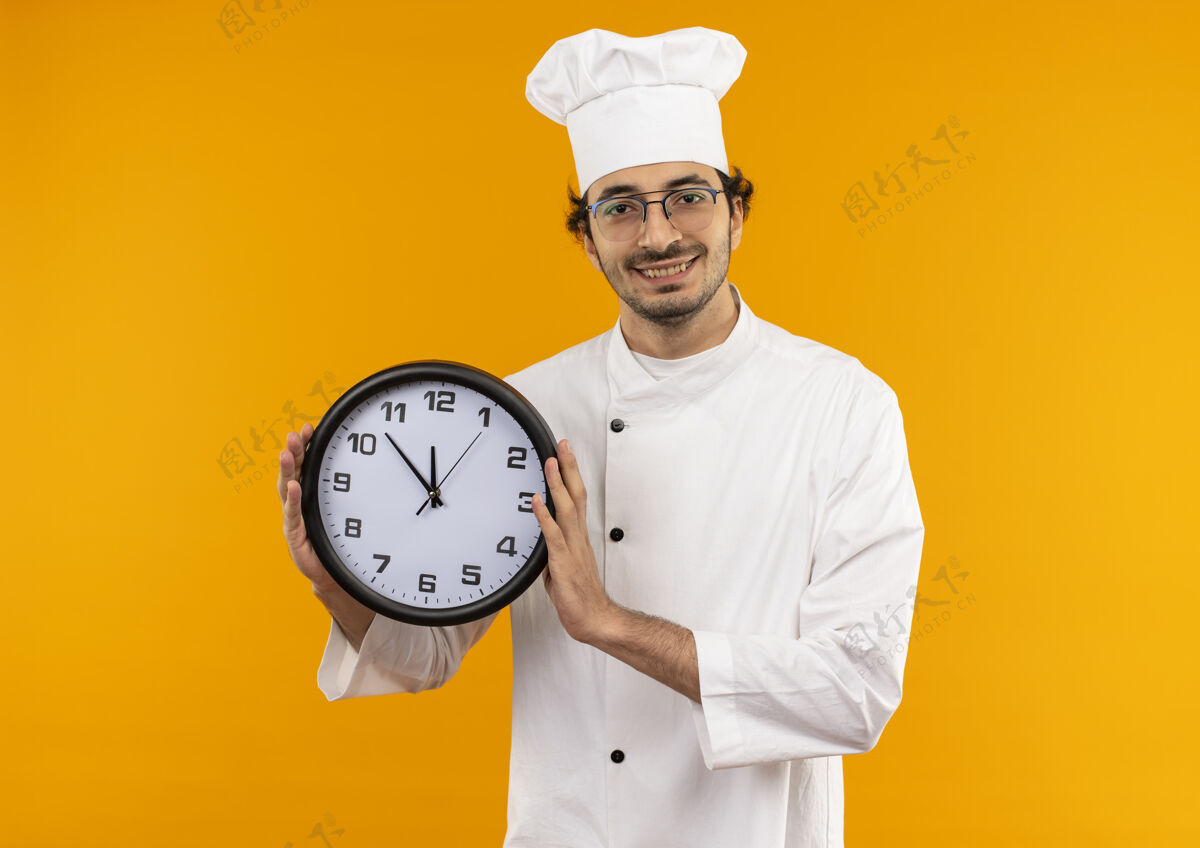 制服面带微笑的年轻男厨师穿着厨师制服 戴着眼镜 把挂钟隔离在黄色的墙上墙壁男性微笑