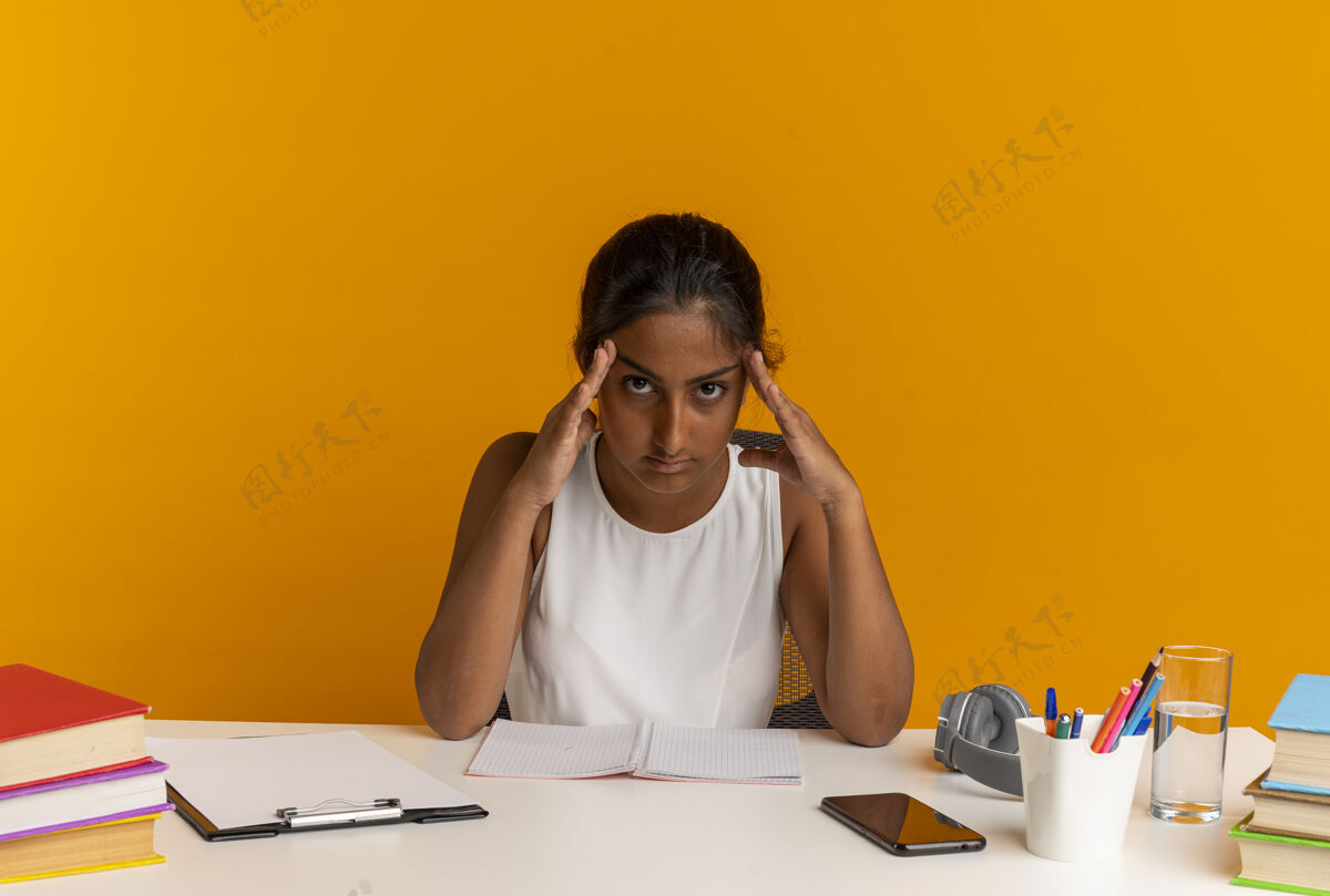 坐着疲惫的年轻女学生坐在课桌旁 手里拿着学习用具 把双手放在孤立在橙色墙上的额头上年轻学校桌子