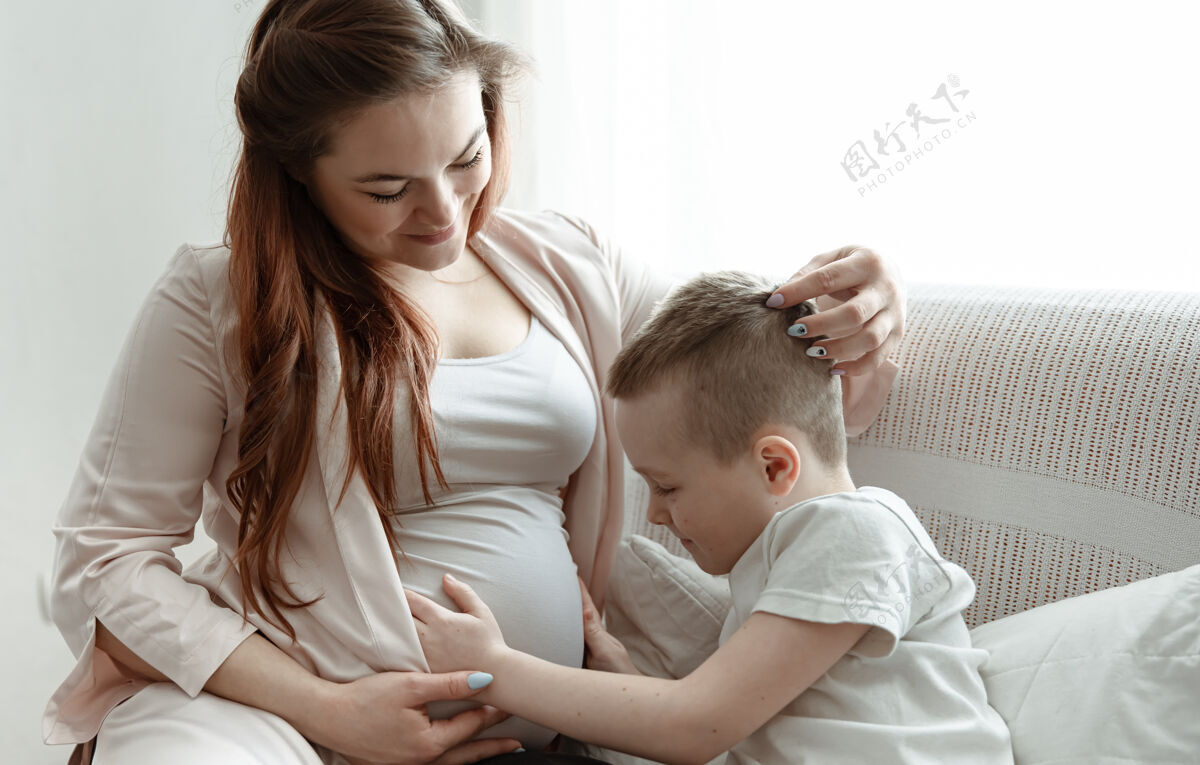 怀孕小男孩在家里的沙发上抱着怀孕母亲的肚子母亲女人父母