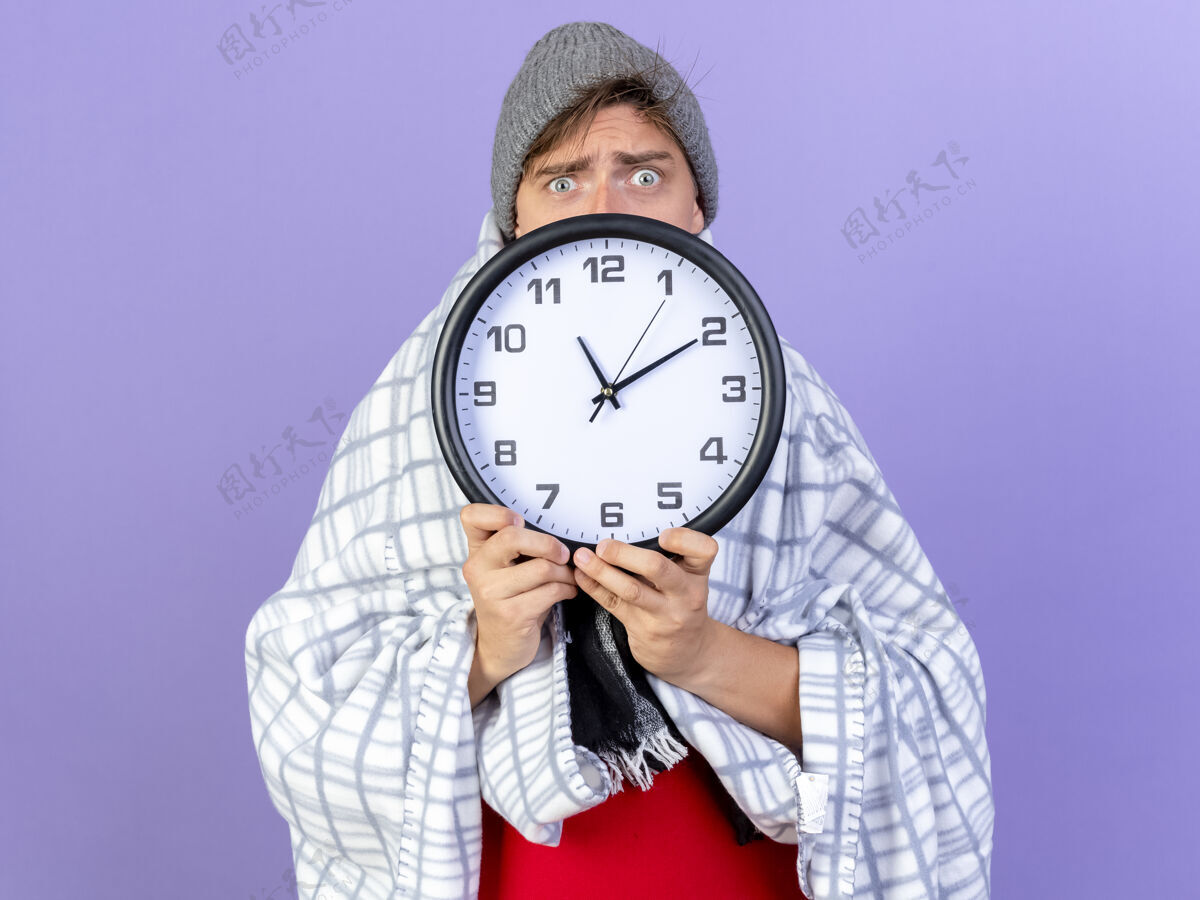 举行印象深刻的年轻英俊的金发病男子戴着冬天的帽子和围巾包裹在格子布拿着时钟看着相机从后面它孤立的紫色背景相机时钟年轻