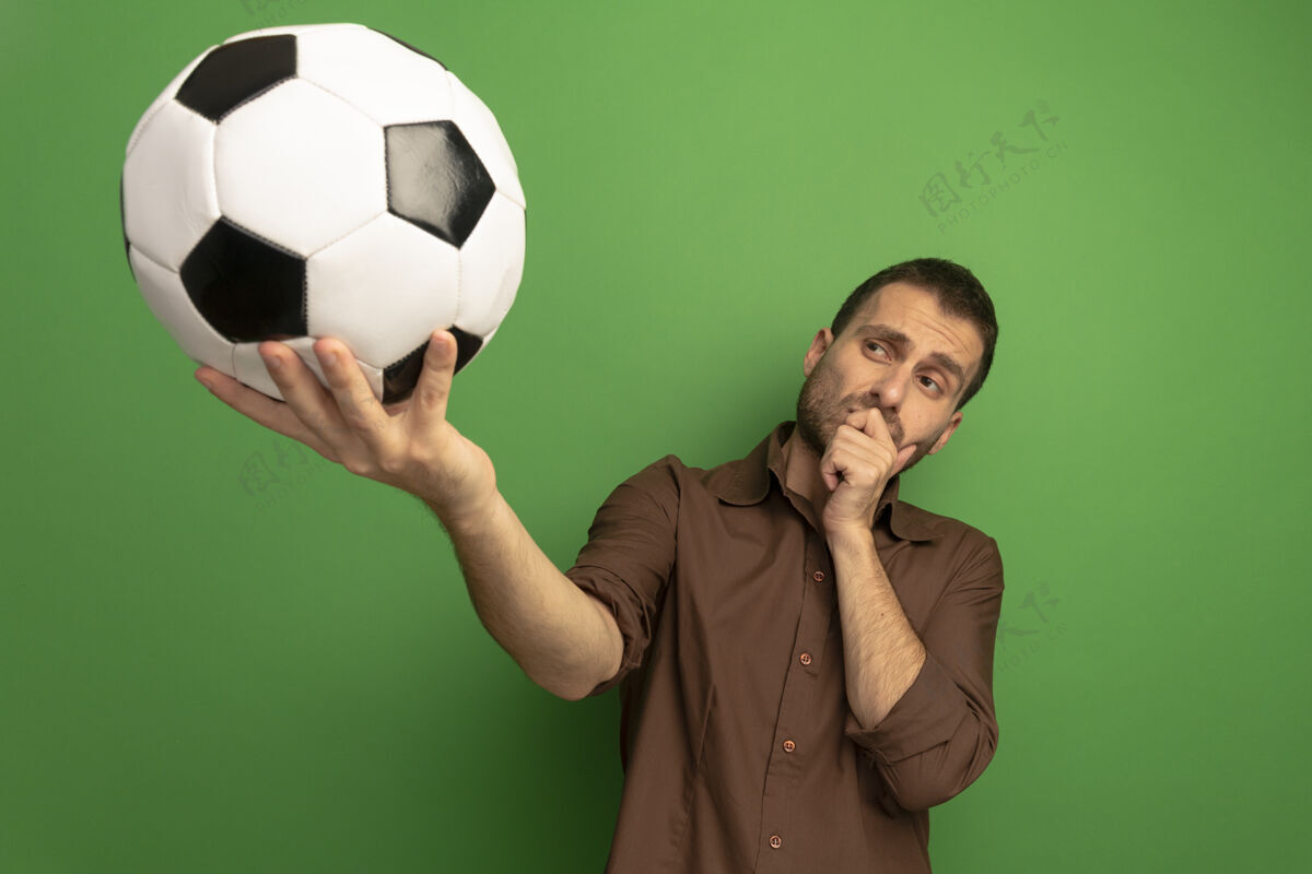 年轻可疑的年轻白种人向镜头伸出足球 手放在下巴上看着球 背景是绿色的 有复印空间朝向相机足球