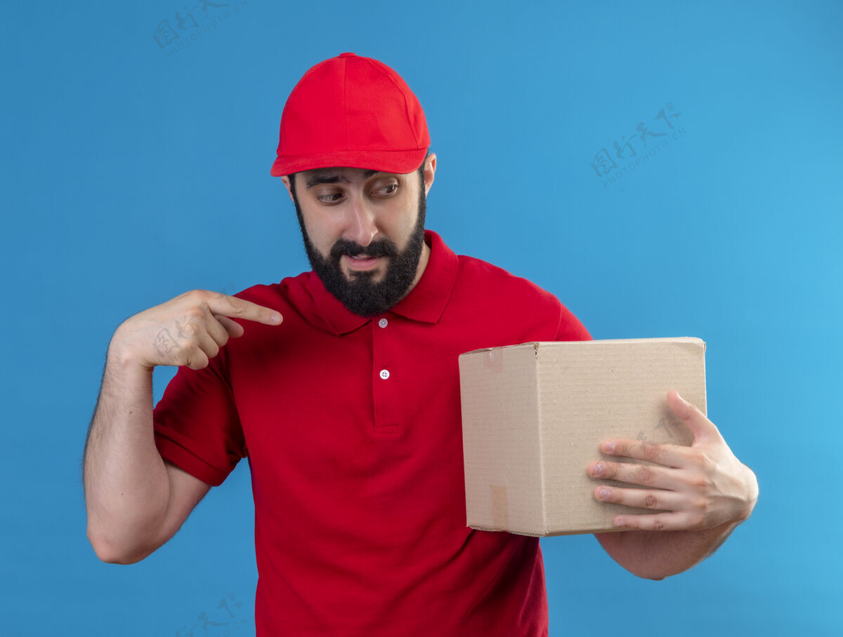 男人印象深刻的年轻英俊的白人送货员穿着红色制服 戴着帽子看着和指着纸箱上孤立的蓝色蓝色指着帅气