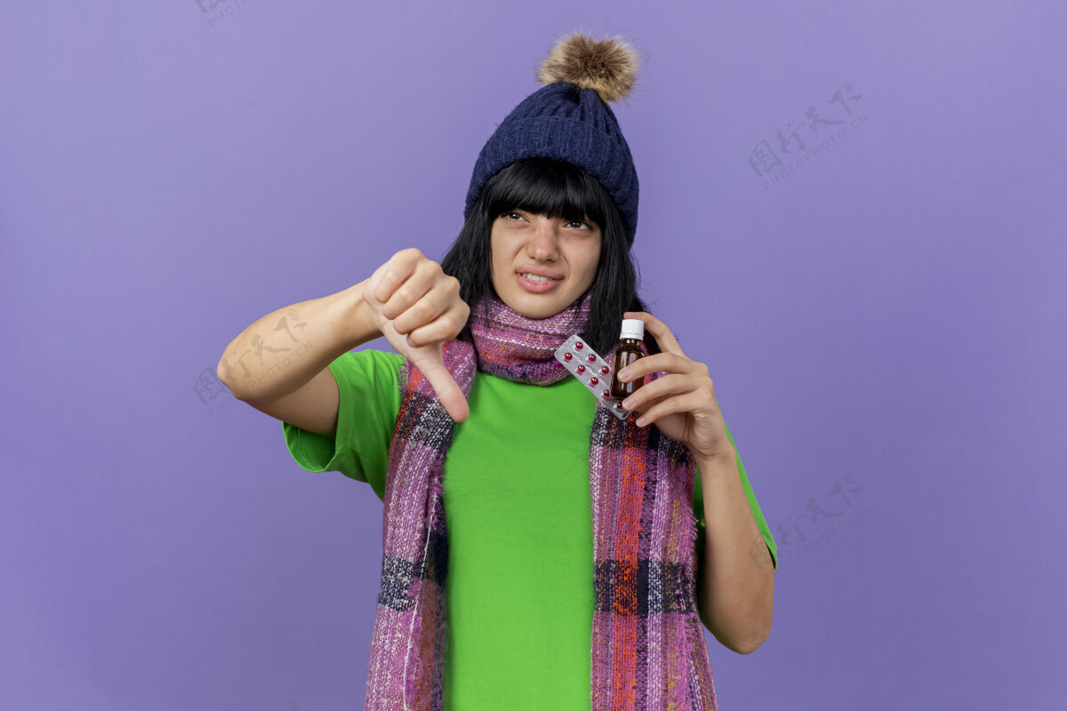 拇指年轻的白种女孩 戴着冬天的帽子 戴着围巾 手里拿着药片 手里拿着一包胶囊 从侧面看 拇指朝下 紫色背景上有一个单独的复制空间不愉快帽子女孩