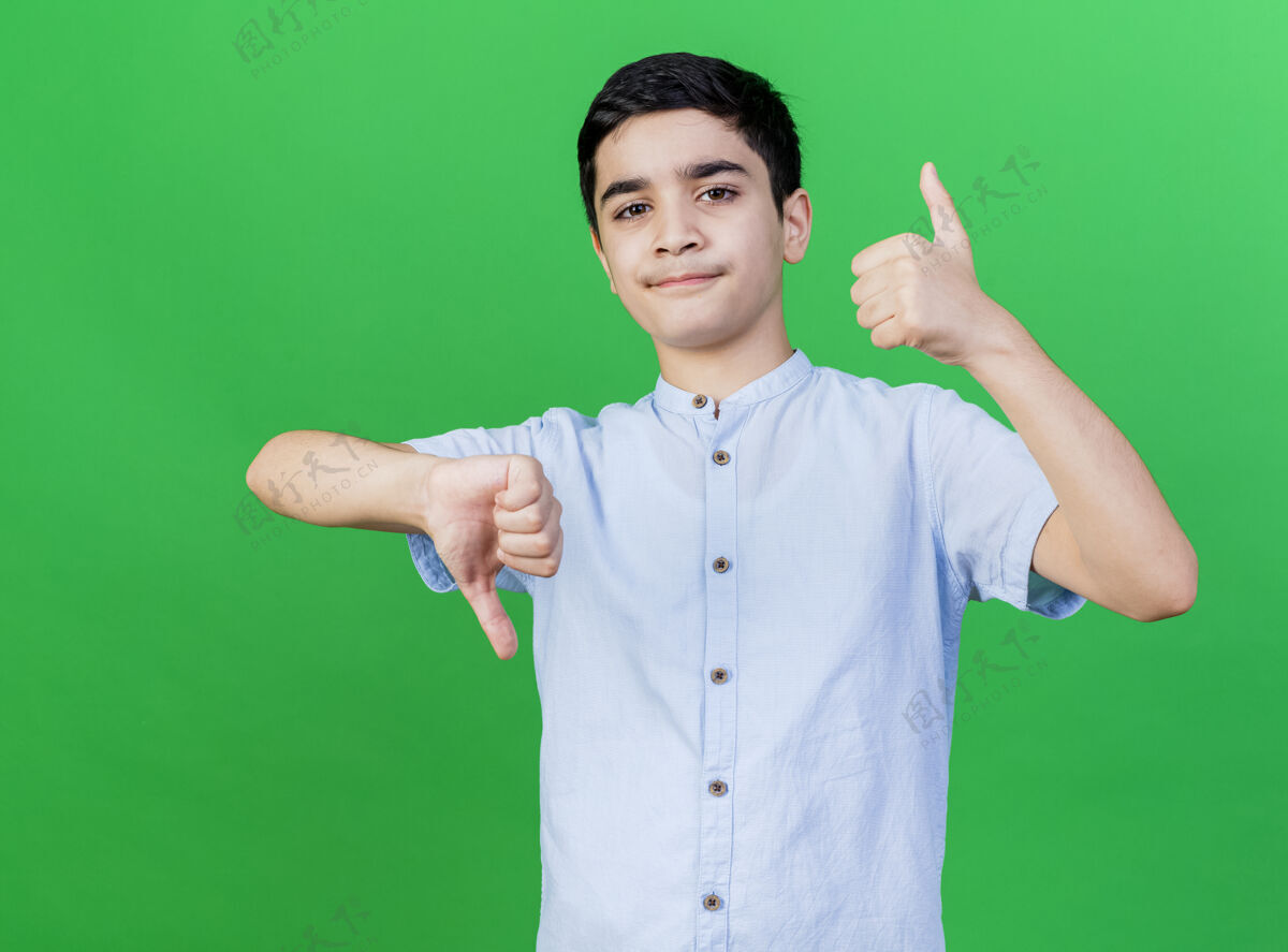年轻一个年轻的白人男孩看着相机 在绿色的背景下上下移动着大拇指令人不快相机男孩