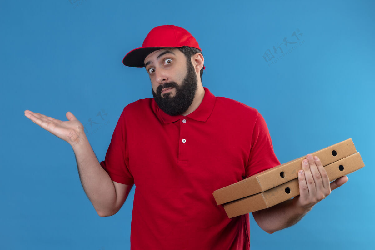 手困惑的年轻英俊的白人送货员身穿红色制服 头戴鸭舌帽 手里拿着比萨饼盒 手上空空如也 孤零零的蓝色帽子迷糊披萨