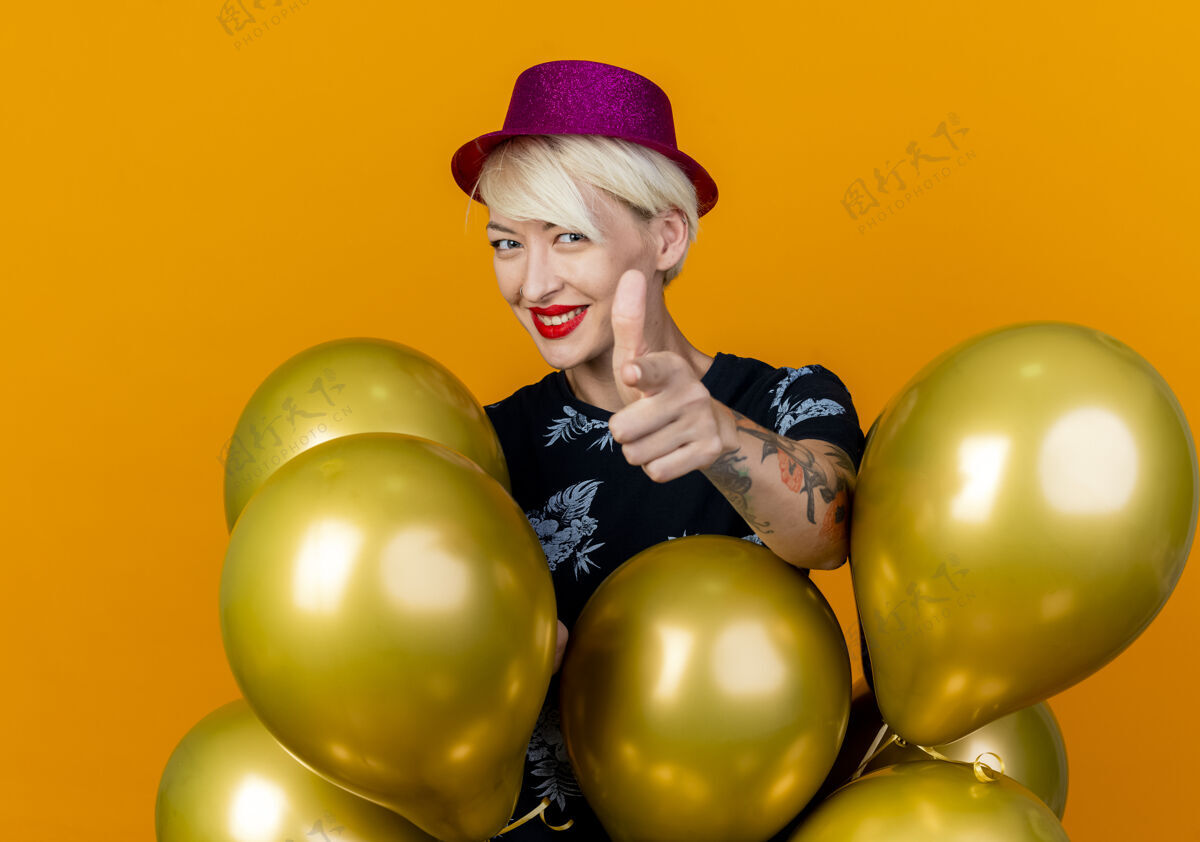 指着微笑着的金发派对女郎 戴着派对帽 站在气球后面 看着和橙色背景隔离的摄像机金发女郎站着年轻人