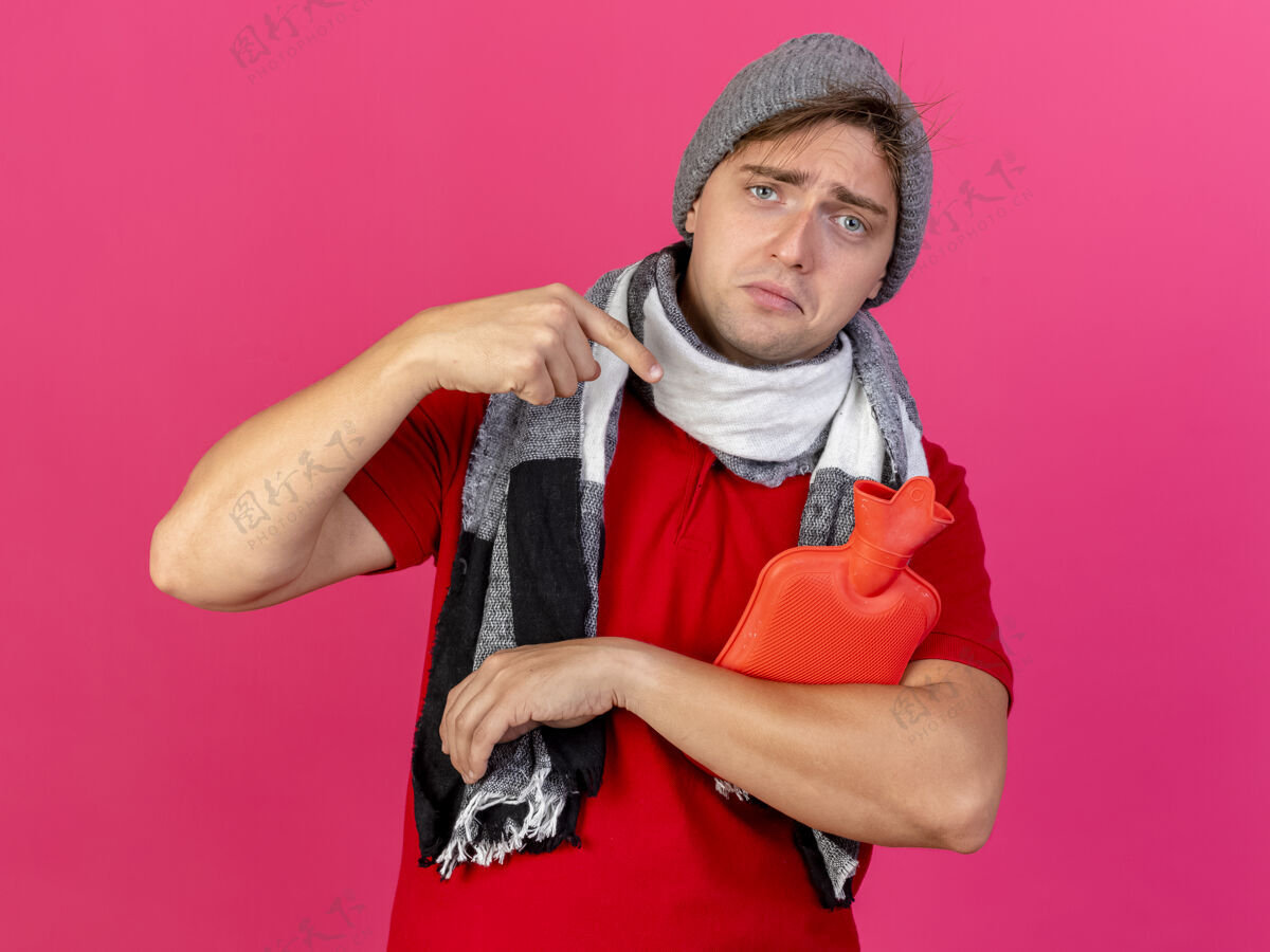 持有悲伤的年轻英俊的金发病男子戴着冬天的帽子和围巾看着相机拿着和指着热水瓶隔离在深红色的背景帽子年轻指着