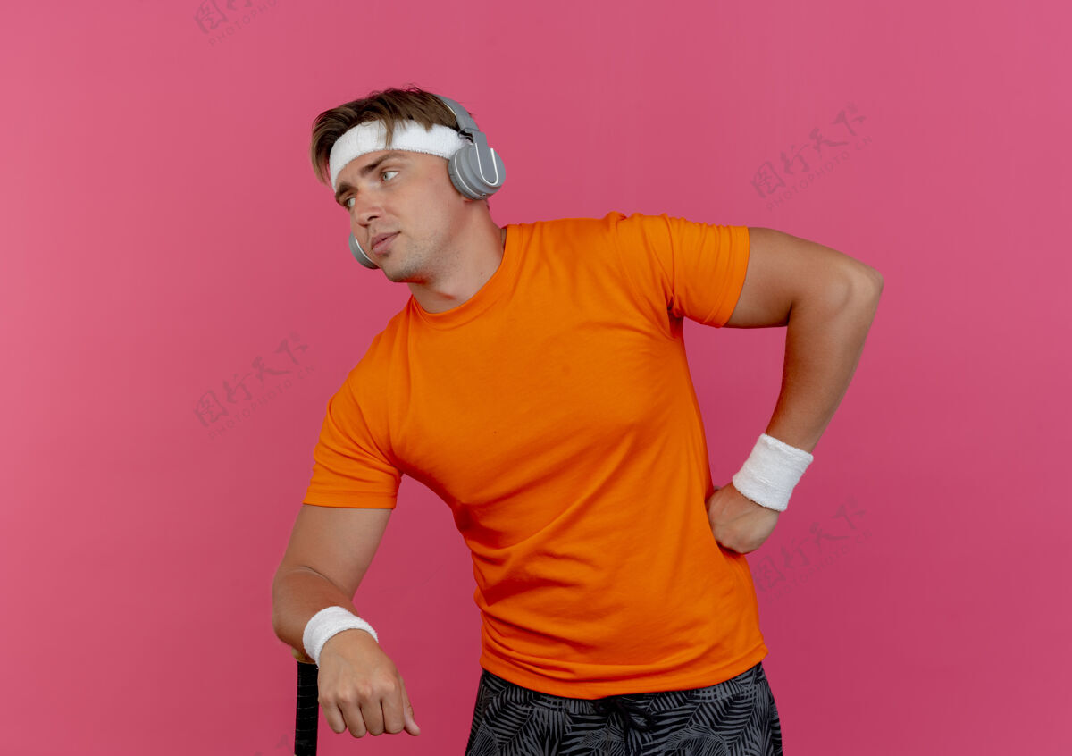 男人自信的年轻帅气的运动型男人戴着头带和腕带 戴着耳机 手放在腰上 另一只手放在棒球棒上 看着粉色的侧面 留着复制空间年轻粉红色头带
