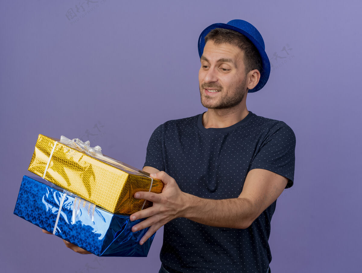 帅哥戴着蓝色帽子的白人帅哥高兴地拿着礼物盒 看着紫色背景上的复制空间盒子帽子请拿着