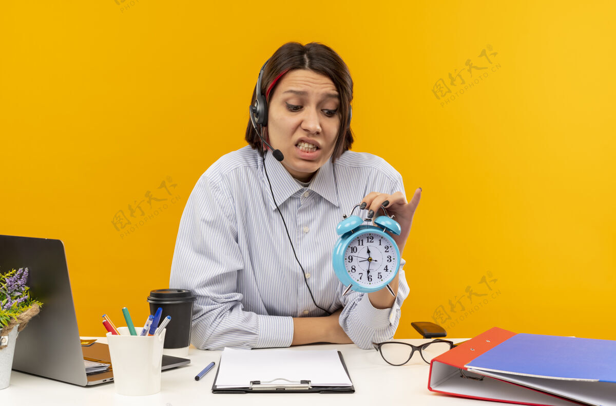 戴着心急如焚的年轻呼叫中心女孩戴着耳机坐在办公桌旁 抱着闹钟看着孤立的桔黄色办公桌焦虑闹钟