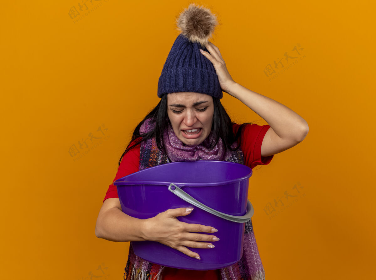 烦躁恼怒和疼痛的年轻白种人生病的女孩戴着冬天的帽子和围巾拿着塑料桶看里面有恶心隔离橙色背景与复制空间水桶年轻女孩