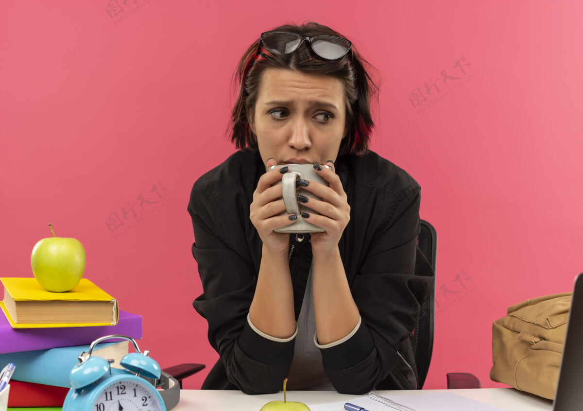 焦虑焦急的年轻女学生头戴眼镜 拿着杯子坐在书桌旁做作业 喝着粉红色的咖啡拿着戴着学生